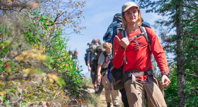 a group of outward bound students hike alone a tree-lined trail with water in the background